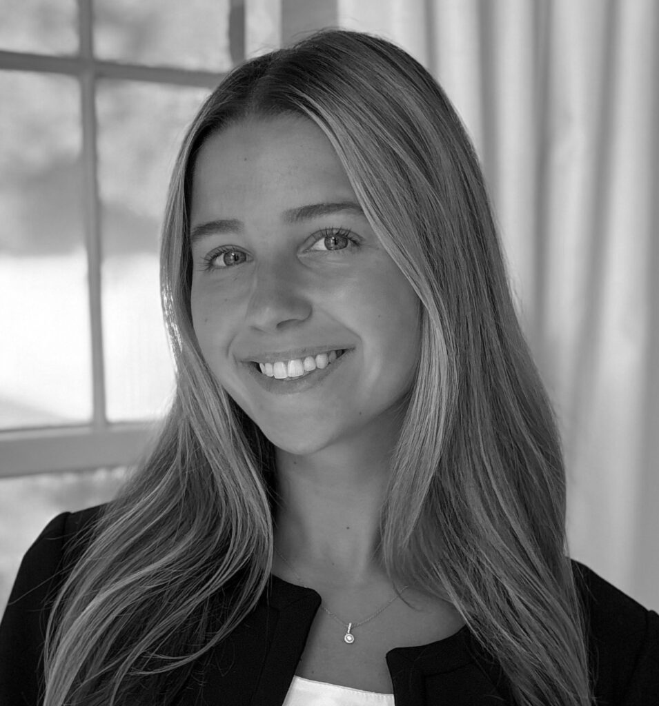 Professional headshot of Kirsten Kizis, a young woman with long straight blonde hair, smiling warmly at the camera, wearing a dark blazer over a light top and a delicate pendant necklace, softly lit near a window, in black and white